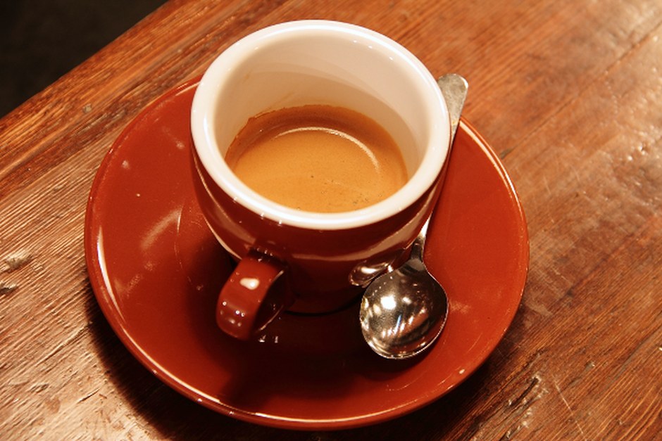 A warm cup of coffee on a red saucer beside a small spoon on a wooden table.