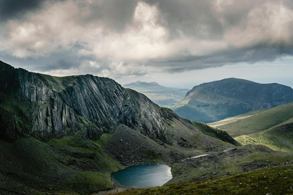 A dramatic green mountain valley with a small lake beneath layered clouds.